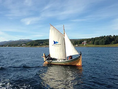 Seilbåt med hvitt seil og blå logo fra Seil og Mesanas, på sjøen nær land.