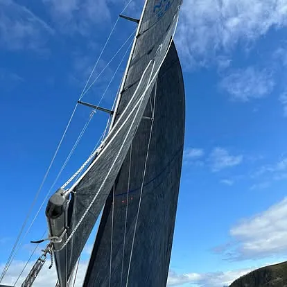 Mørkt seil i full høyde på båt under seilas med blå himmel i bakgrunnen.