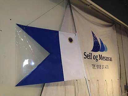 Blå og hvit vimpel med emblem, hengt opp foran banner med logoen "Seil og Mesanas".