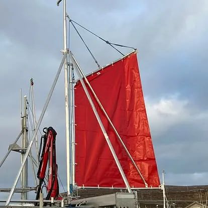 Rødt firkantseil montert i mast på båt ved kai, med grå himmel bak.