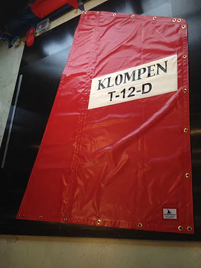 Rød presenning med teksten "KLOMPEN T-12-D" og maljer langs kanten.
