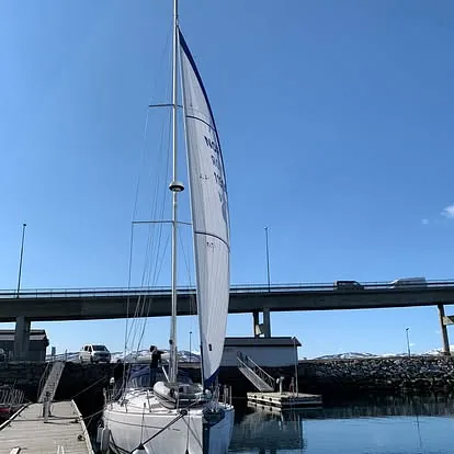 Seilbåt med heist hvitt forseil ligger fortøyd ved brygge under klar blå himmel.